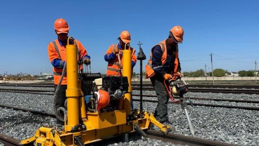 Three rail workers wearing high-visibility orange safety gear and hard hats operate equipment on a set of railway tracks.